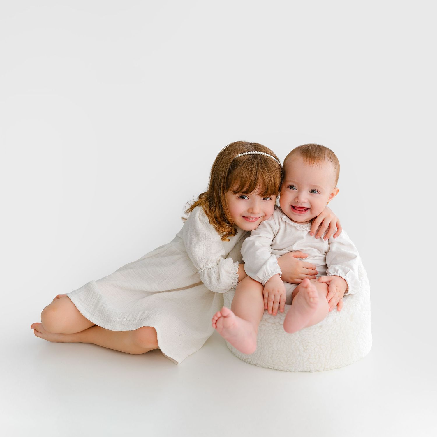 Sweet studio portrait of siblings in white outfits sitting together against white background.