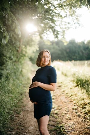 Pregnant woman in navy blue dress holding belly on sunlit nature trail maternity portrait.