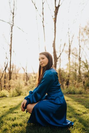Young woman in blue satin dress sitting on grass in a sunlit woodland setting.