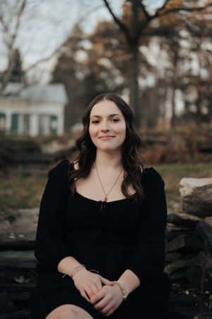Young woman in black dress sitting on stone fence smiling softly in an autumn outdoor setting.