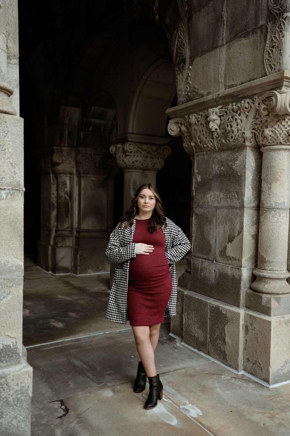 Pregnant woman in red dress and houndstooth coat poses gracefully between ornate stone columns.