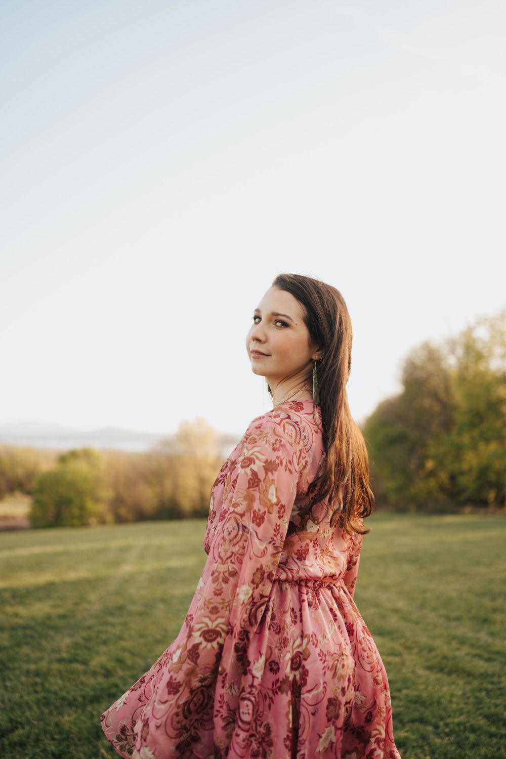 Woman in floral pink dress twirling on green hillside at golden hour with scenic landscape.