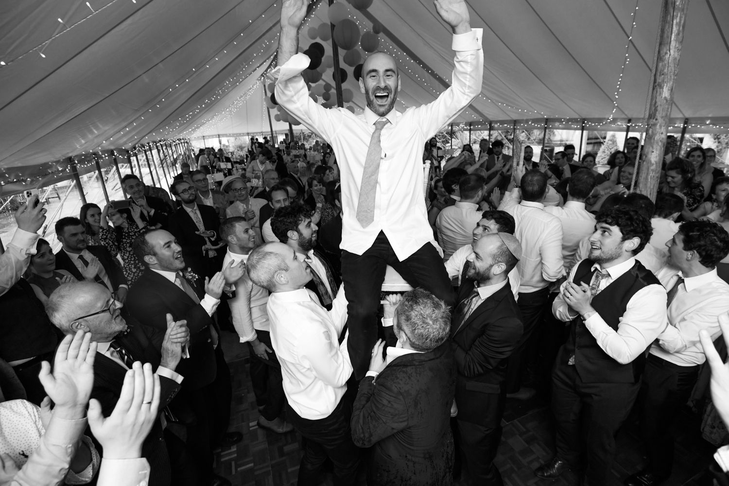 Groom lifted by excited wedding guests during celebration under tent.