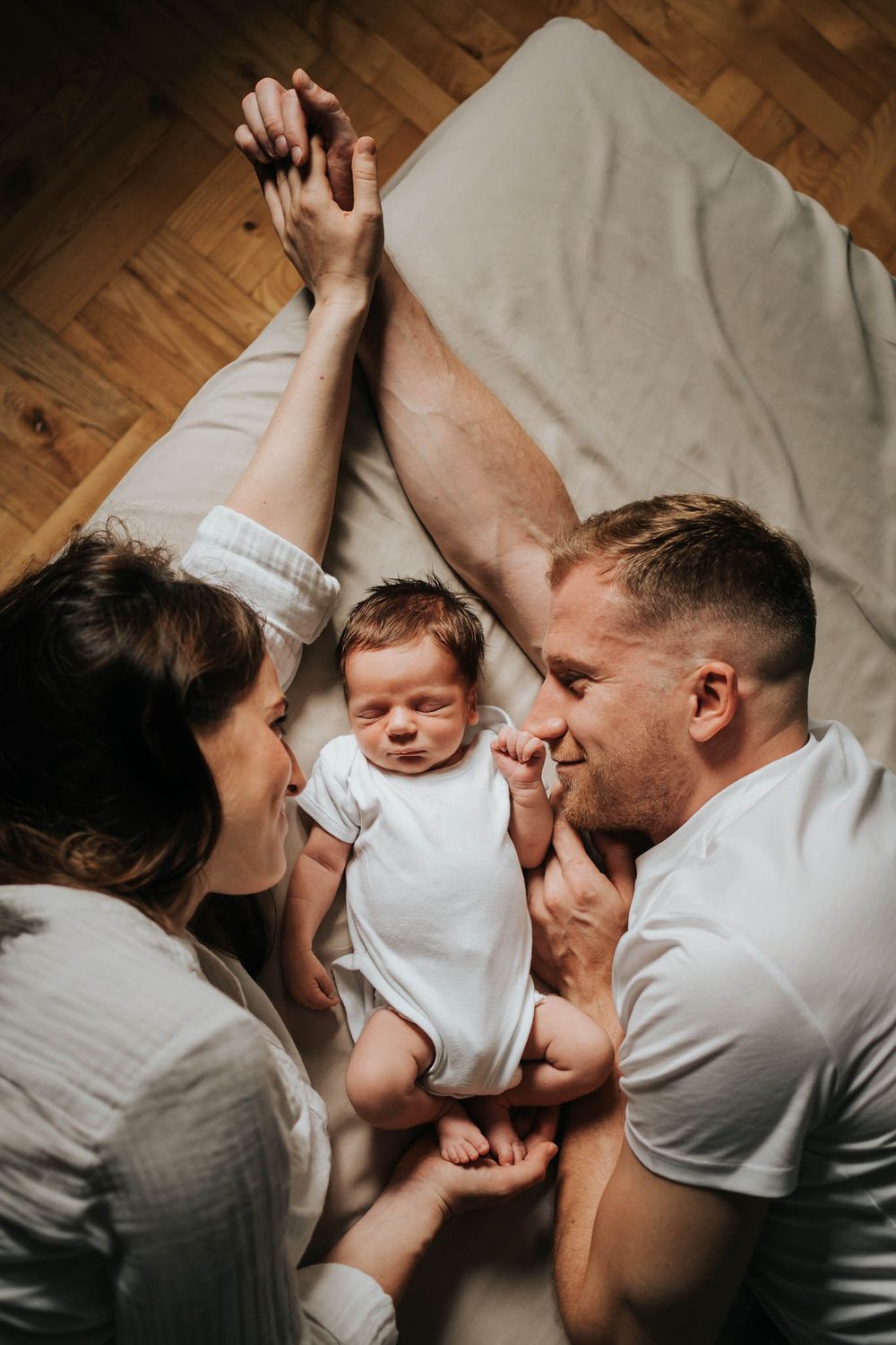 mama i tata patrzą na siebie z miłością, trzymają się za ręce, między nimi leży ich mały synek - sesja noworodkowa Kraków