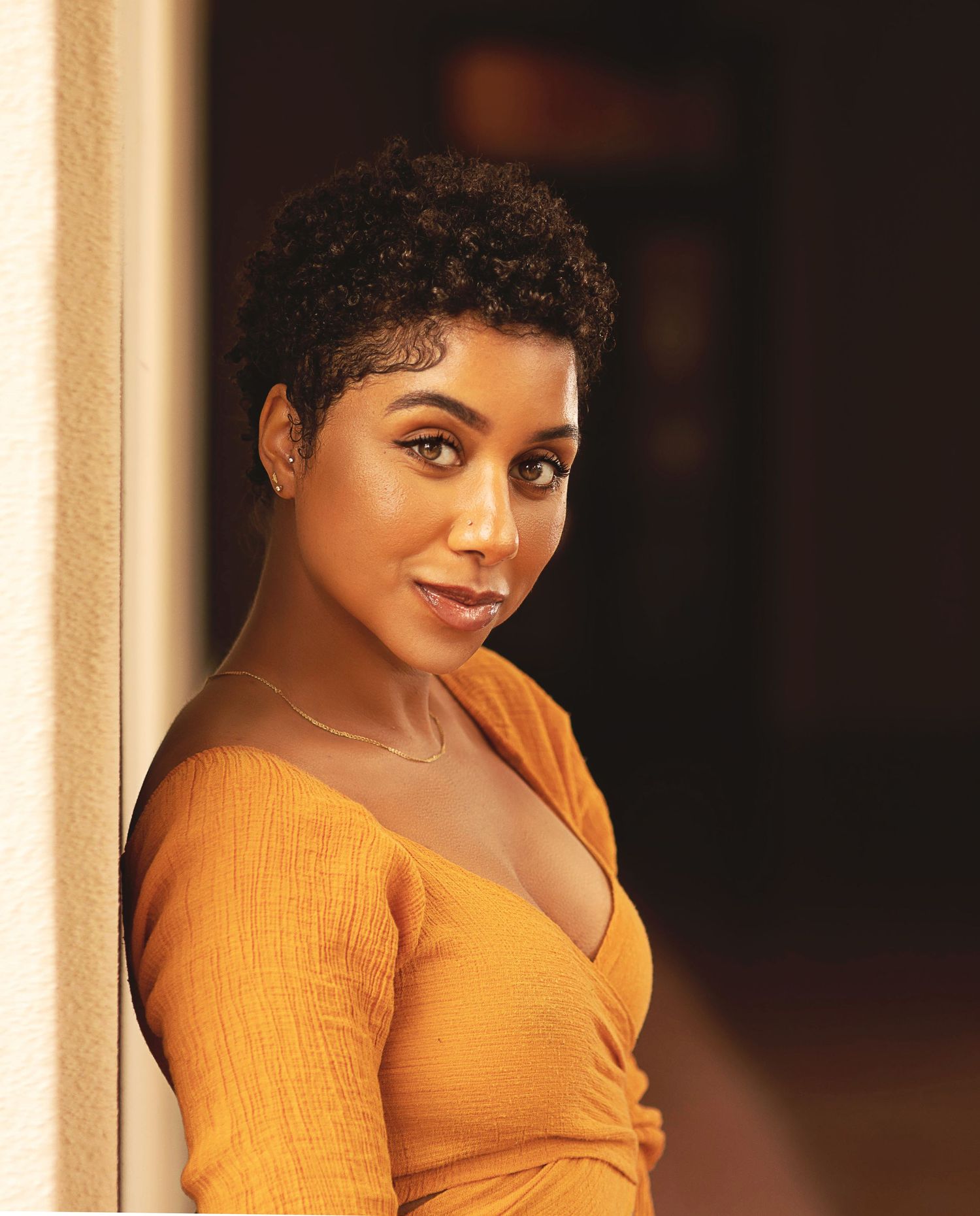 Portrait of woman in orange top leaning against wall with natural lighting and soft focus.