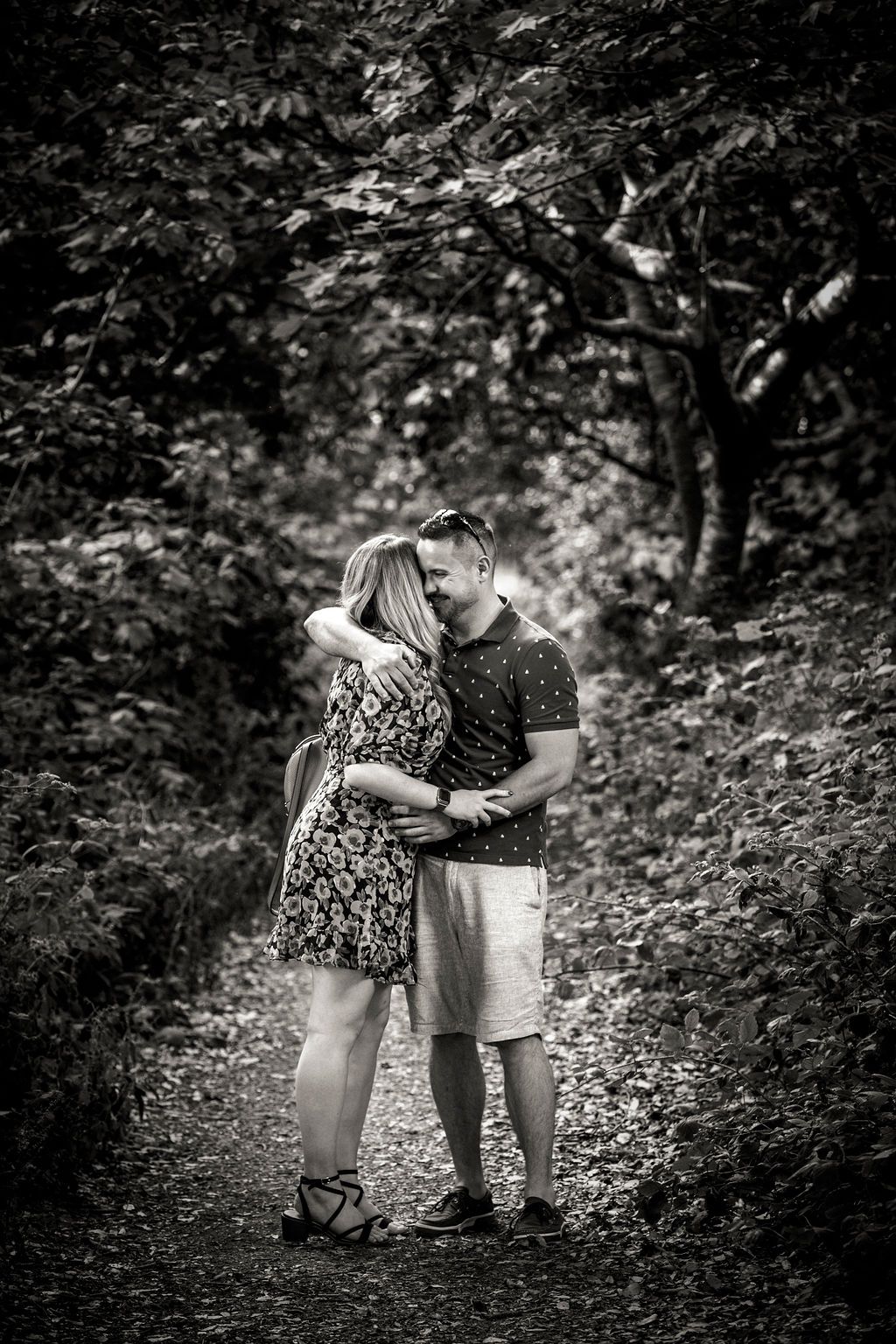 A romantic black and white portrait of a couple embracing on a wooded path.