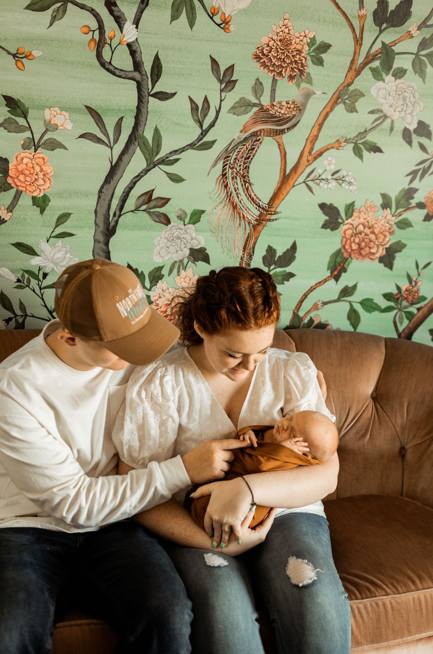 Couple with newborn baby sitting on couch against floral wallpaper.