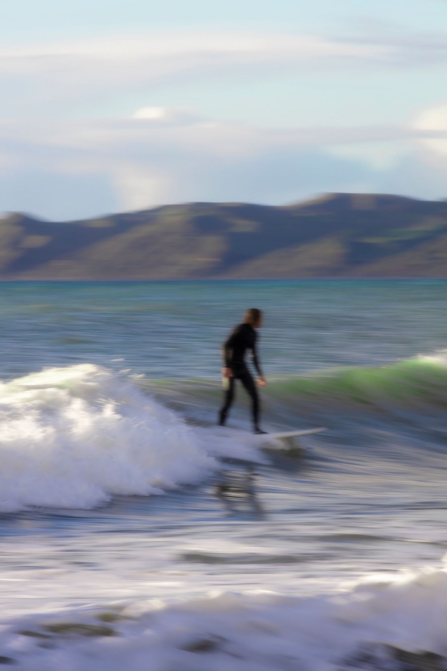 A lone surfer rides a gentle wave near the shoreline with golden light reflecting off the water. Rolling hills form the backdrop under a sof