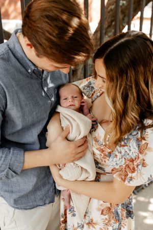 A couple in casual clothing tenderly holds their newborn baby wrapped in white against an outdoor backdrop.