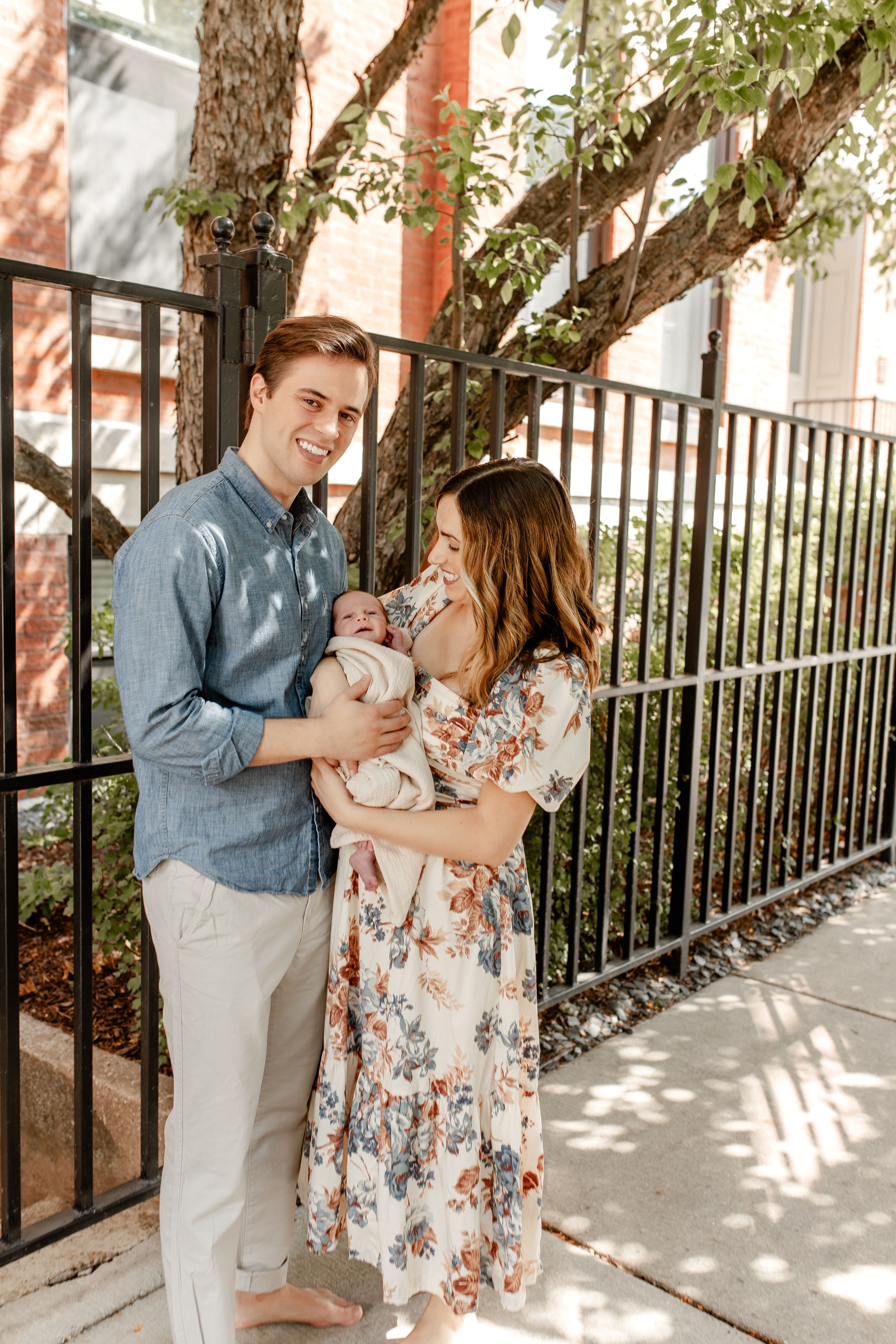 Family portrait outdoors with newborn baby against iron fence and trees.