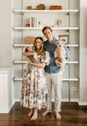 A couple holds their small dogs while standing barefoot in front of white shelves in their home.