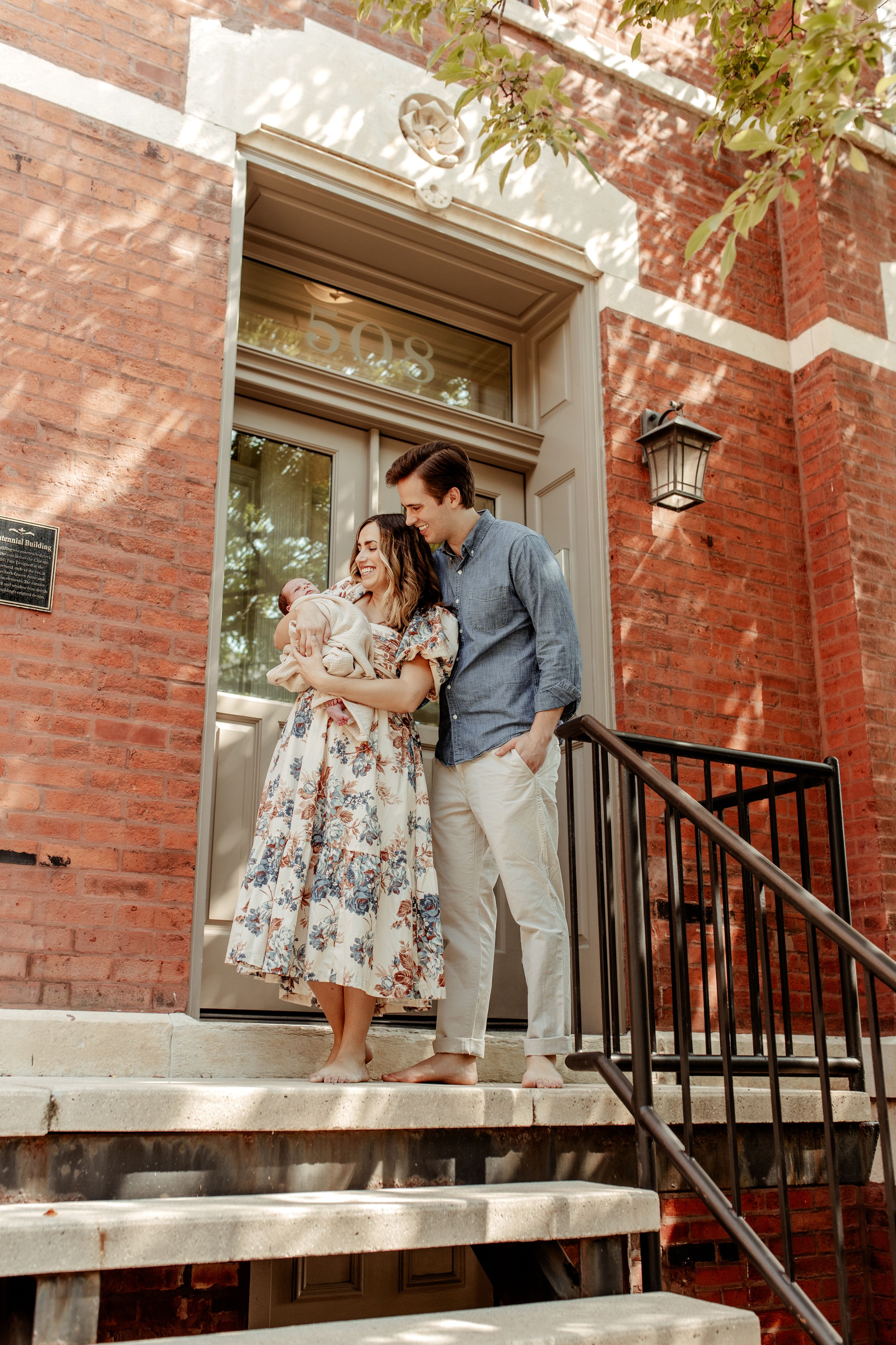 A couple stands on brick building steps holding their baby on a sunny day.