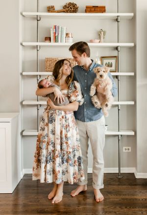 A couple poses with their pets and baby in front of decorative white shelves in their home.