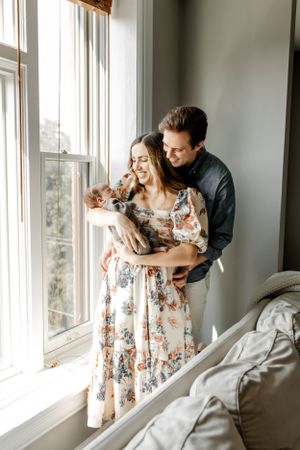 A couple shares a tender moment by a bright window while holding their small dogs.