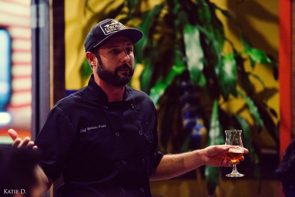 A bearded chef in a black uniform holds a glass of craft beer at a brewery tasting event.