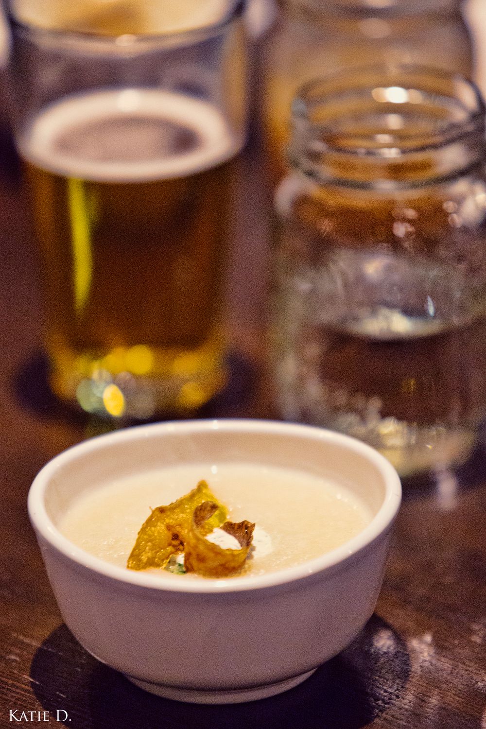 A small white bowl of creamy soup with a crispy garnish beside glasses of craft beer.