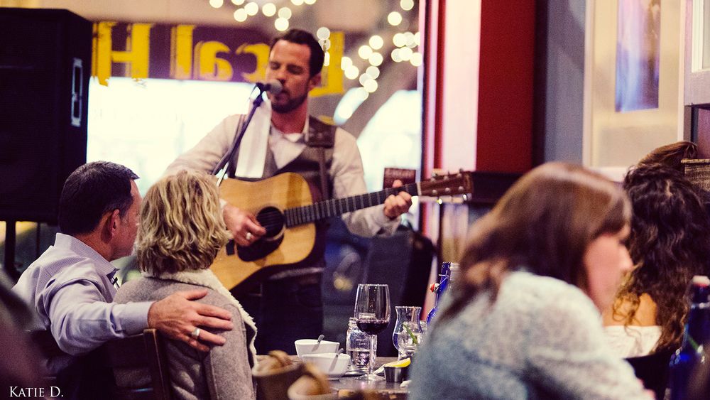 A live musician performs acoustic guitar while restaurant guests enjoy their meals nearby.