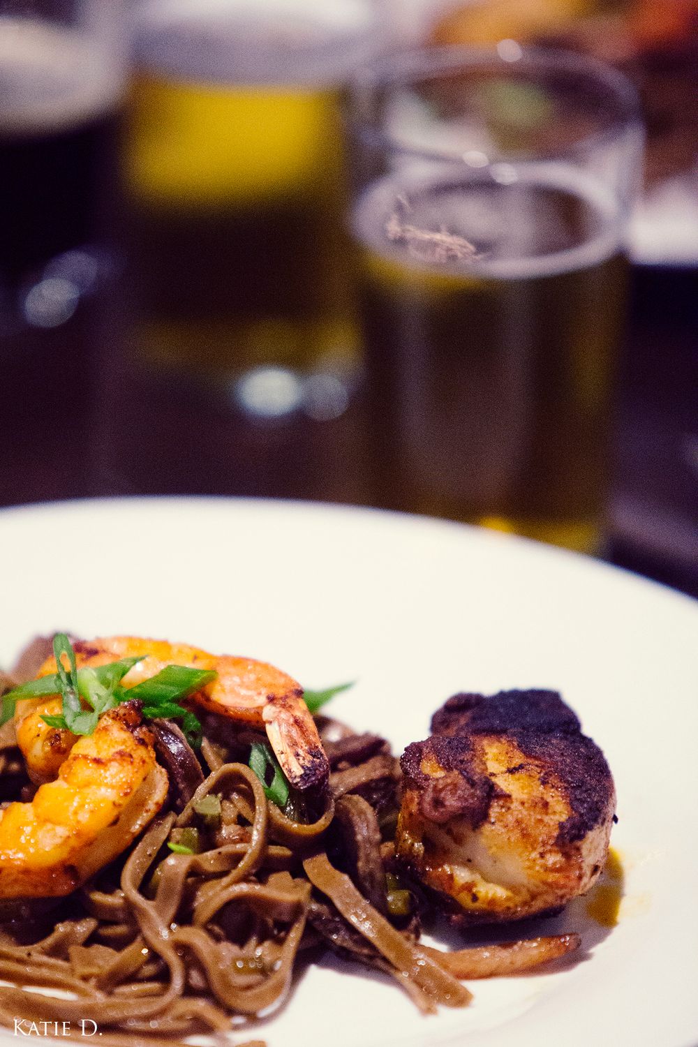 A gourmet plate of seared scallops and shrimp over dark noodles paired with craft beer.