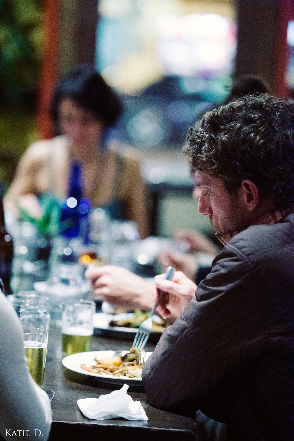 Man with curly hair dining at a restaurant table, holding a fork over a plate of food.