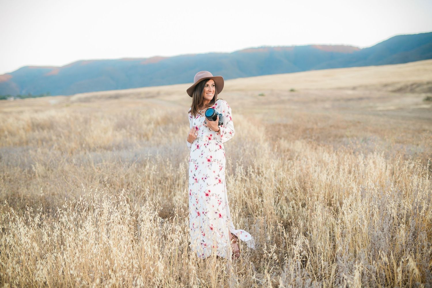Woman in floral maxi dress and hat holding a camera while standing in a golden meadow with mountains.