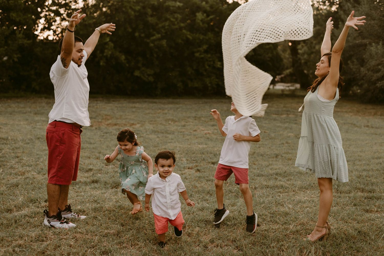 Family jumps joyfully while playing with white fabric in golden evening light.