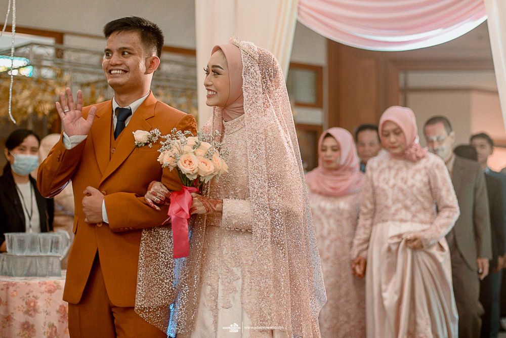A couple in traditional Muslim wedding attire, with the groom wearing an orange suit and bride in pink lace hijab.
