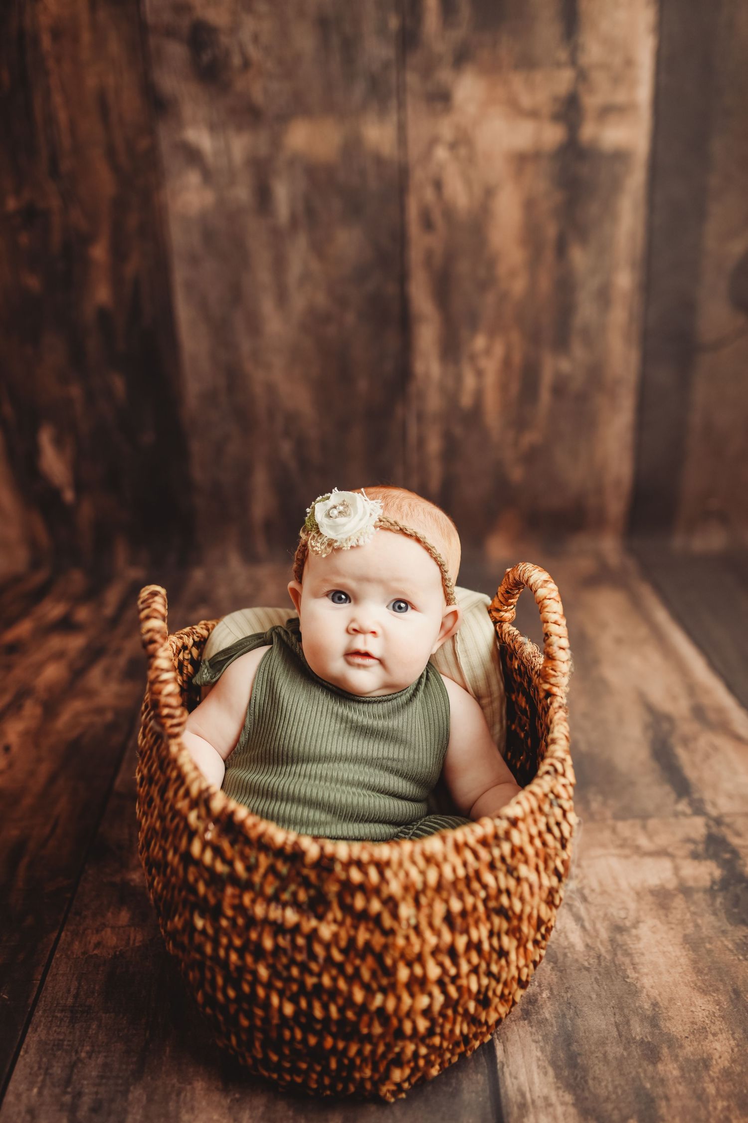 Baby in olive green outfit nestled in textured brown basket against rustic wooden background.