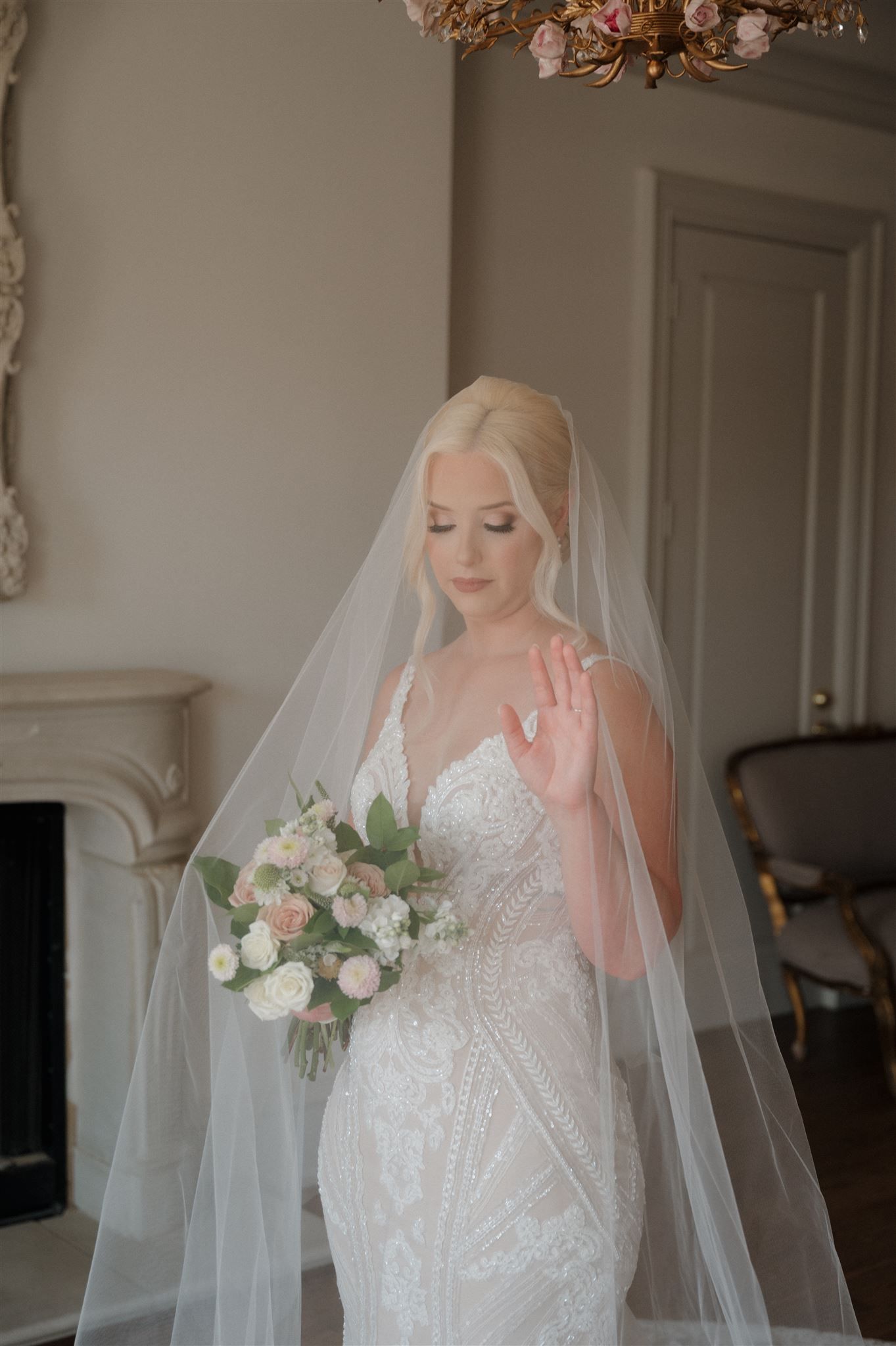 Elegant bride in lace wedding gown holds white and blush bouquet while posing near fireplace in natural light.