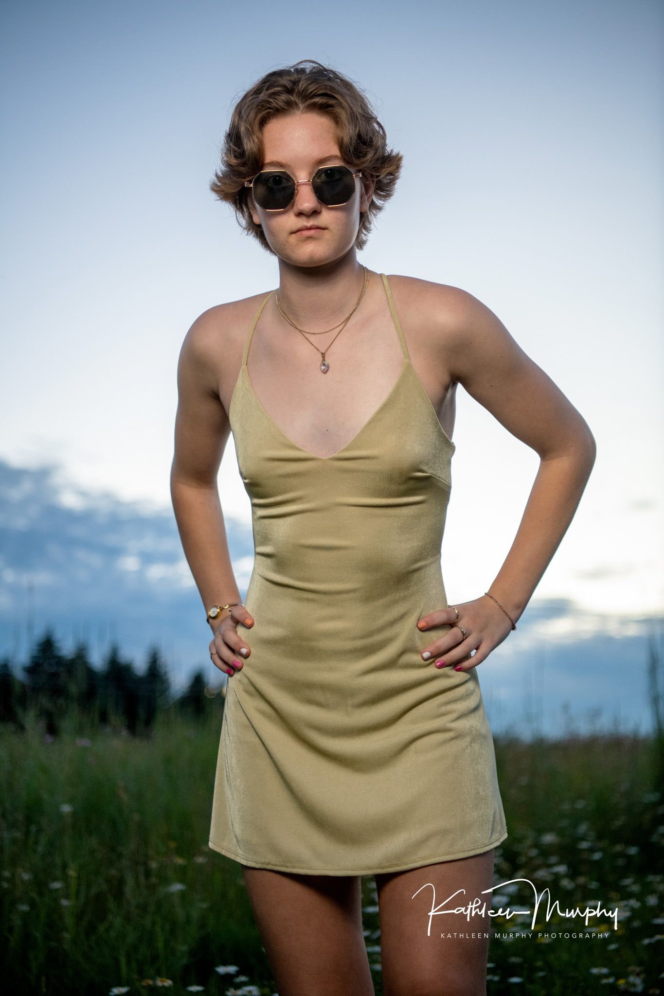 Fashion portrait of someone in a beige slip dress and sunglasses against a mountain backdrop at sunset.