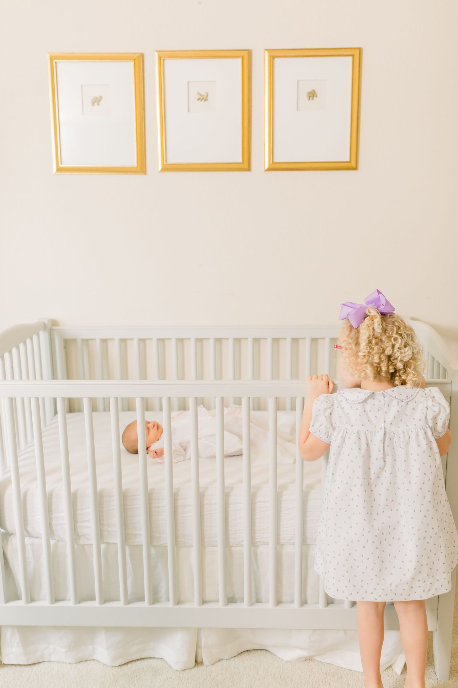 A small child in a white dress looks into a white crib beneath three framed prints.