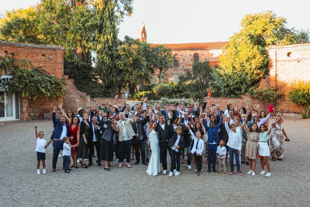 Large group wedding photo with guests cheering and celebrating in courtyard.