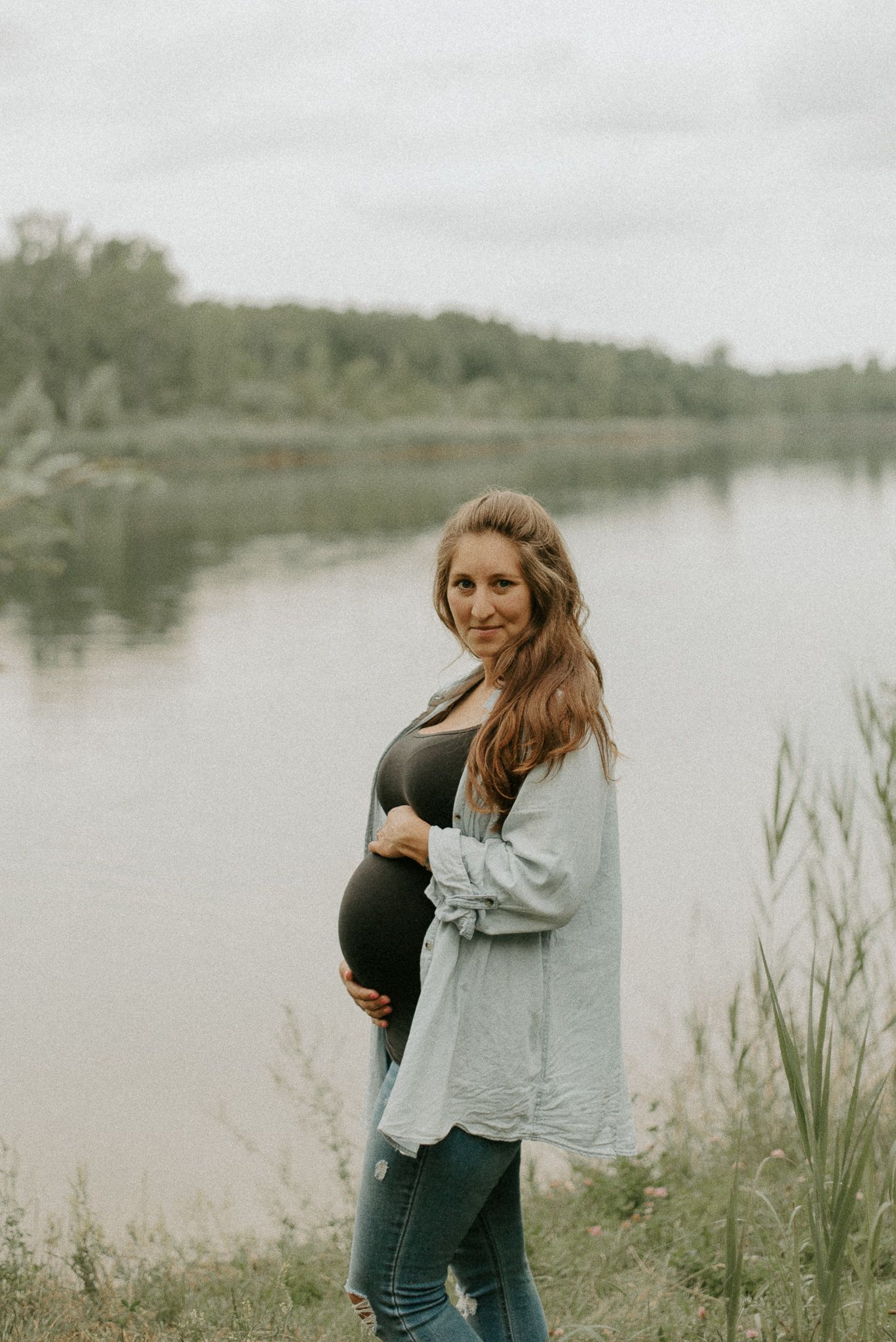 Maternity portrait by serene lake with woman in casual attire.