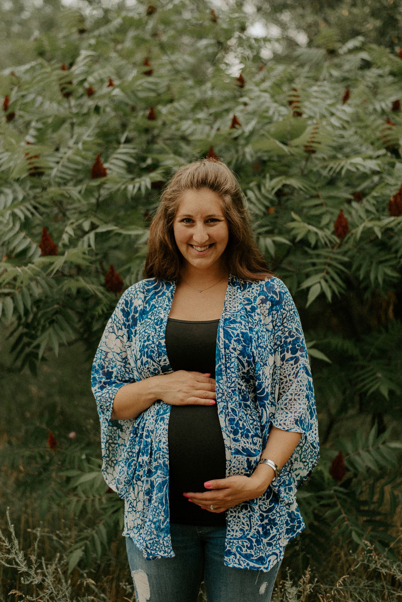 Maternity photo of a woman in a blue floral kimono standing in front of green summer foliage.