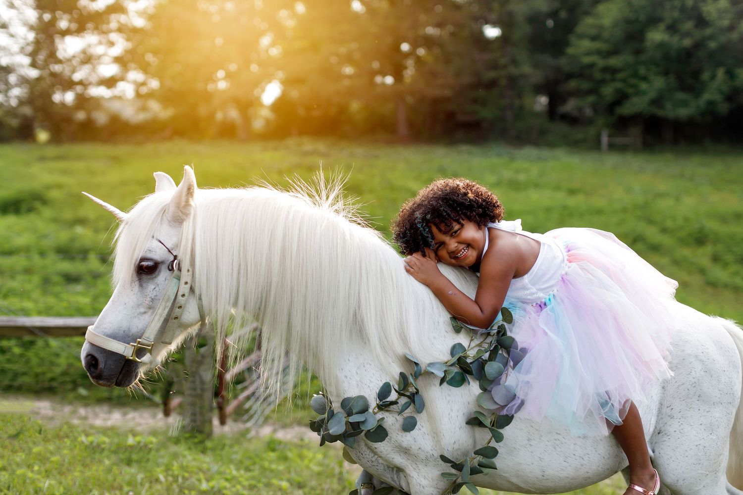 A white pony adorned with greenery stands in a sunlit meadow while someone in a flowing white dress leans against it.