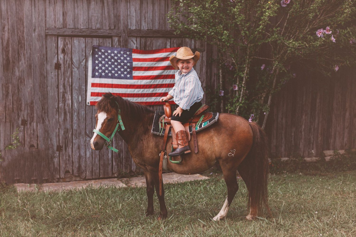 A person in a cowboy hat sits atop a brown horse beside a rustic barn with an American flag hanging behind them.