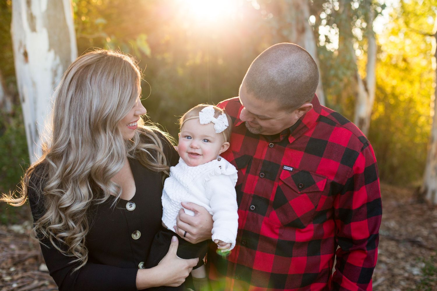A family shares a tender moment in the golden sunlight among trees with a baby in white and dad wearing buffalo plaid.