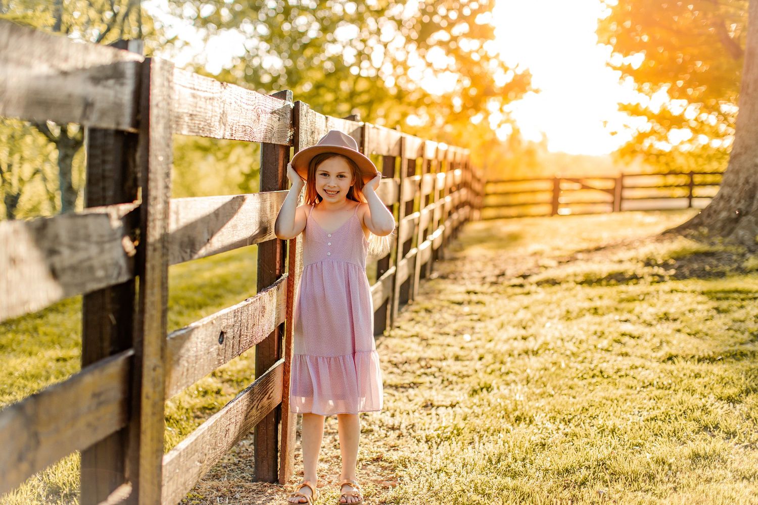 Young girl in pink dress standing by wooden fence during sunset.