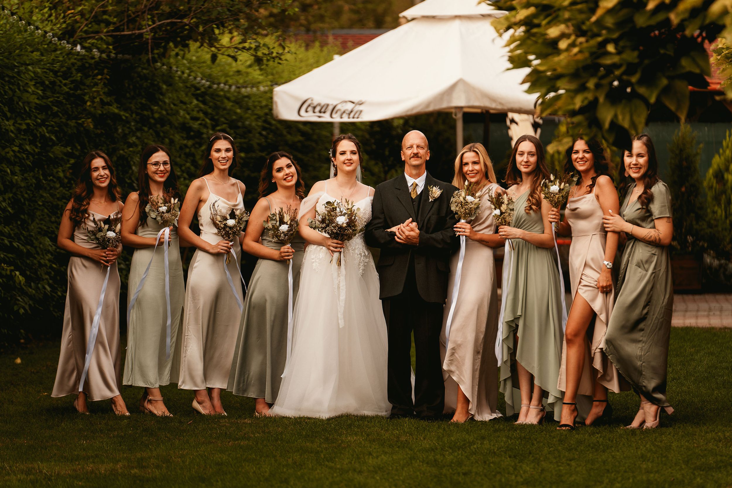 A wedding party poses for a formal group photo outdoors with bridesmaids in matching dresses.