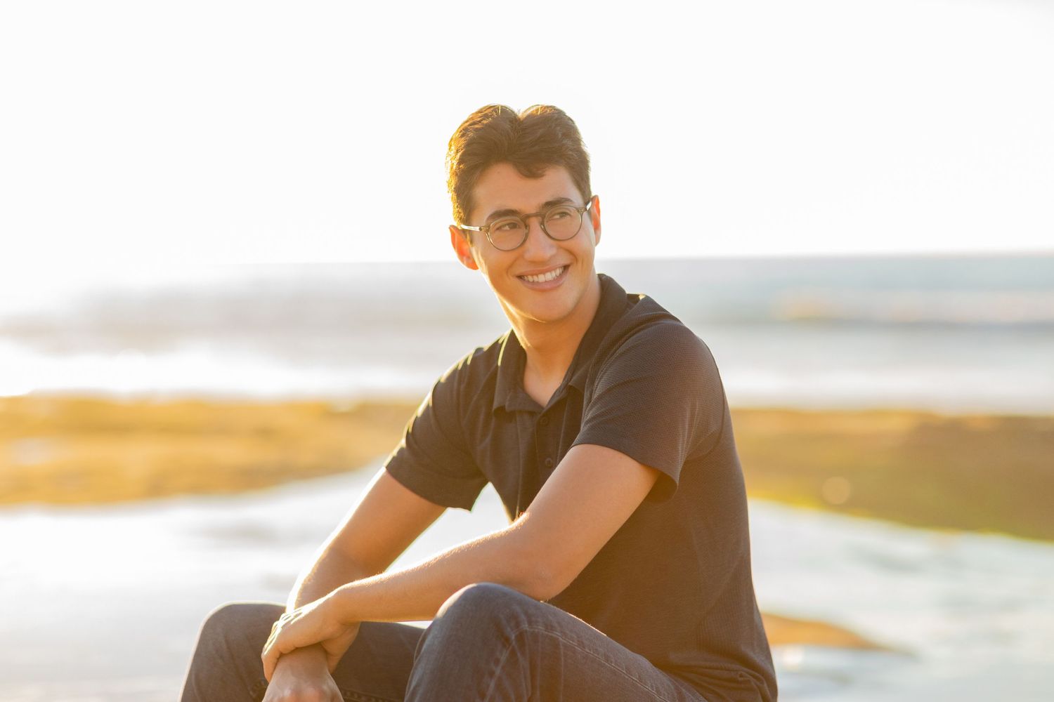 A high school senior in black polo shirt sitting by water during golden hour sunset.