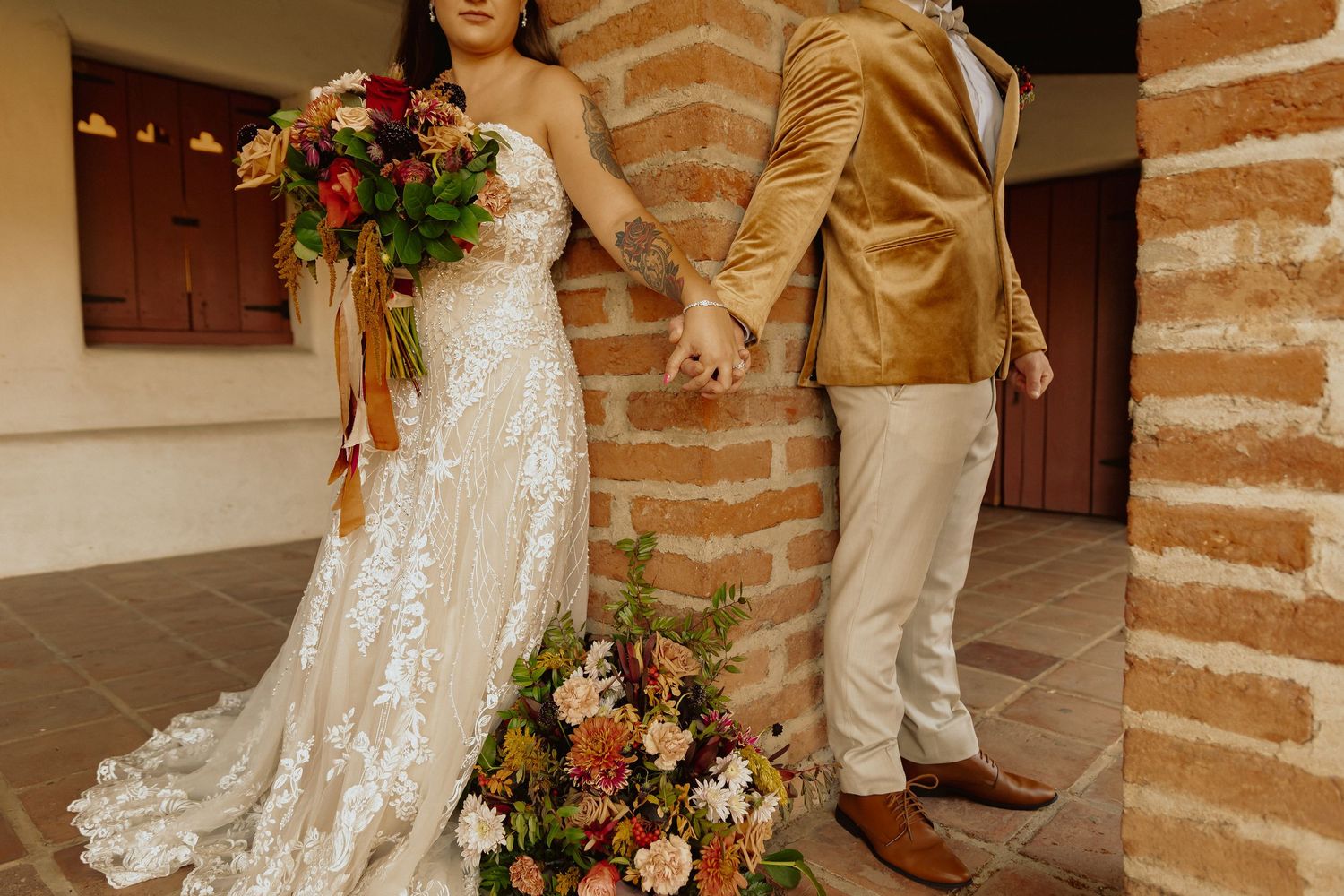 Bride in lace gown holding vibrant bouquet next to groom against brick wall.