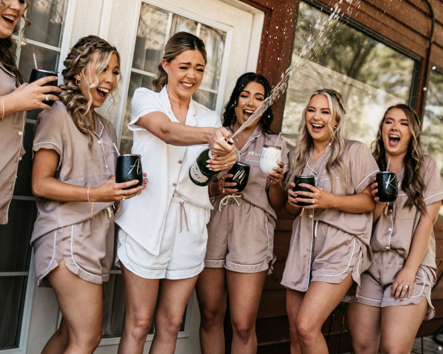 Friends in matching sleepwear celebrate with champagne and laughter on a porch.