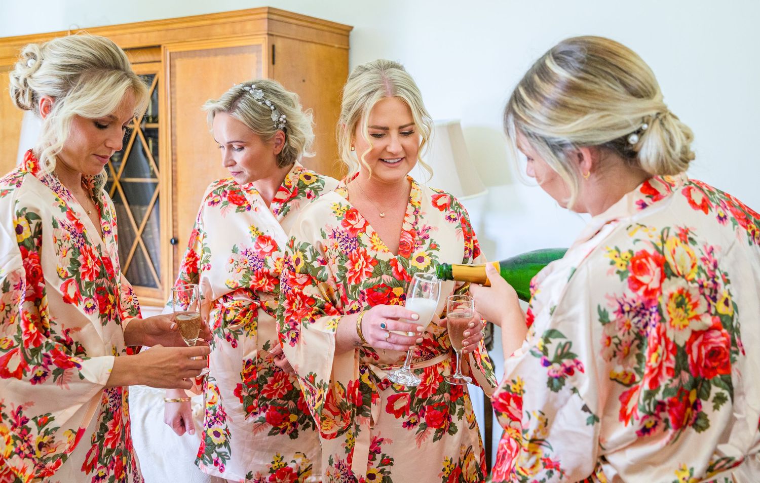 Group wearing matching floral robes toasting with champagne during wedding preparations.