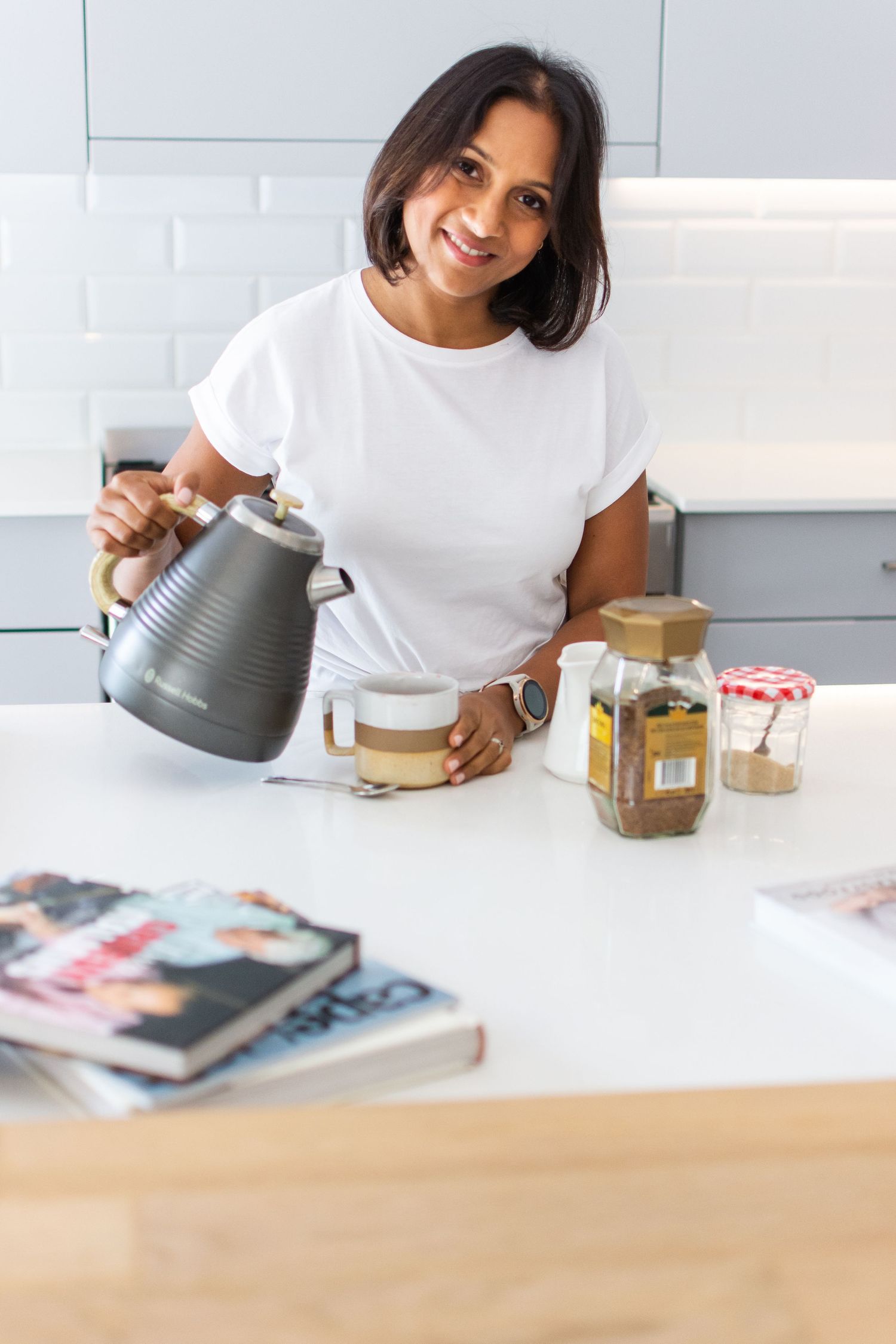Woman pouring hot beverage from kettle in modern white kitchen with cookbooks.