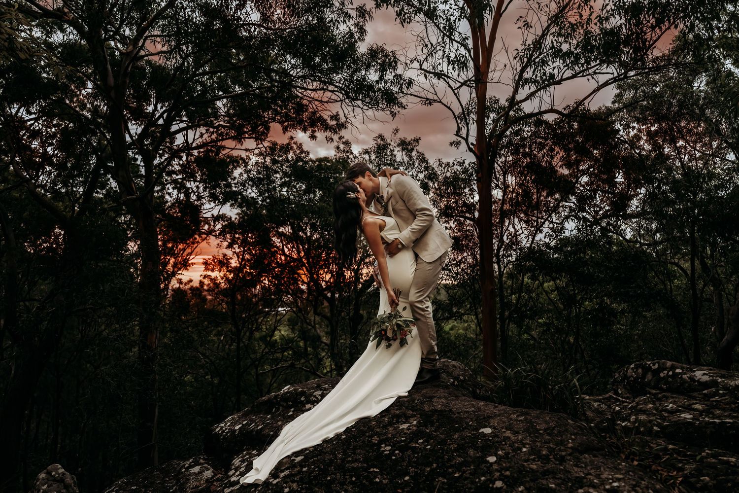 Artistic silhouette of wedding couple against sunset through trees.
