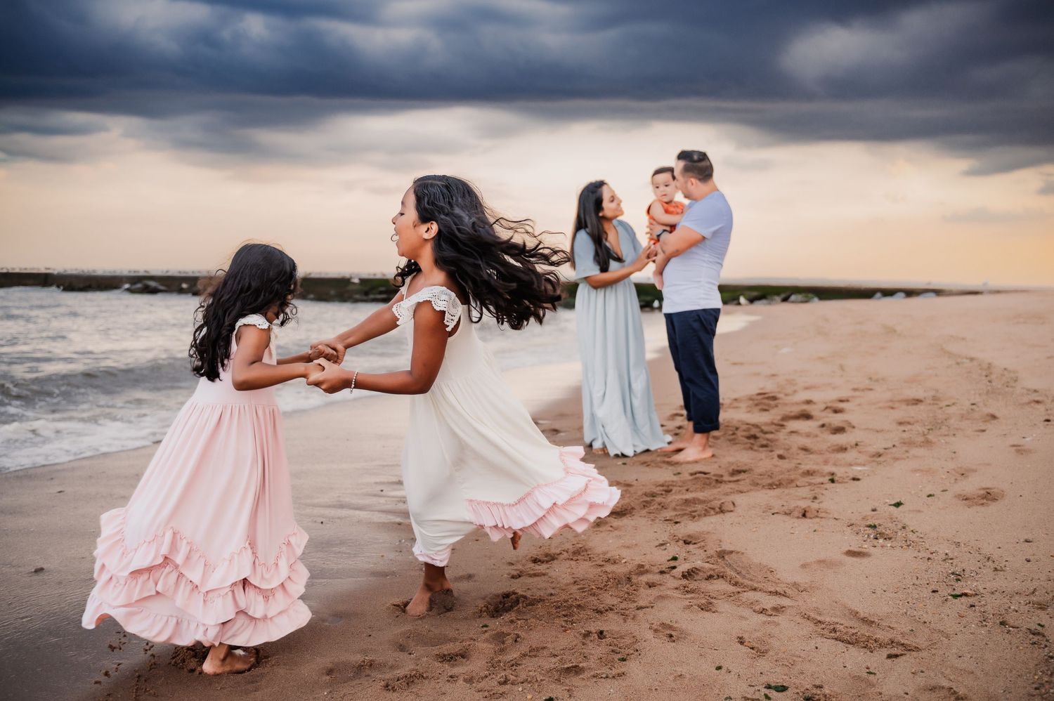 Two young girls holding hands and twirling in front of mother and father holding baby on South Beach in Staten Island