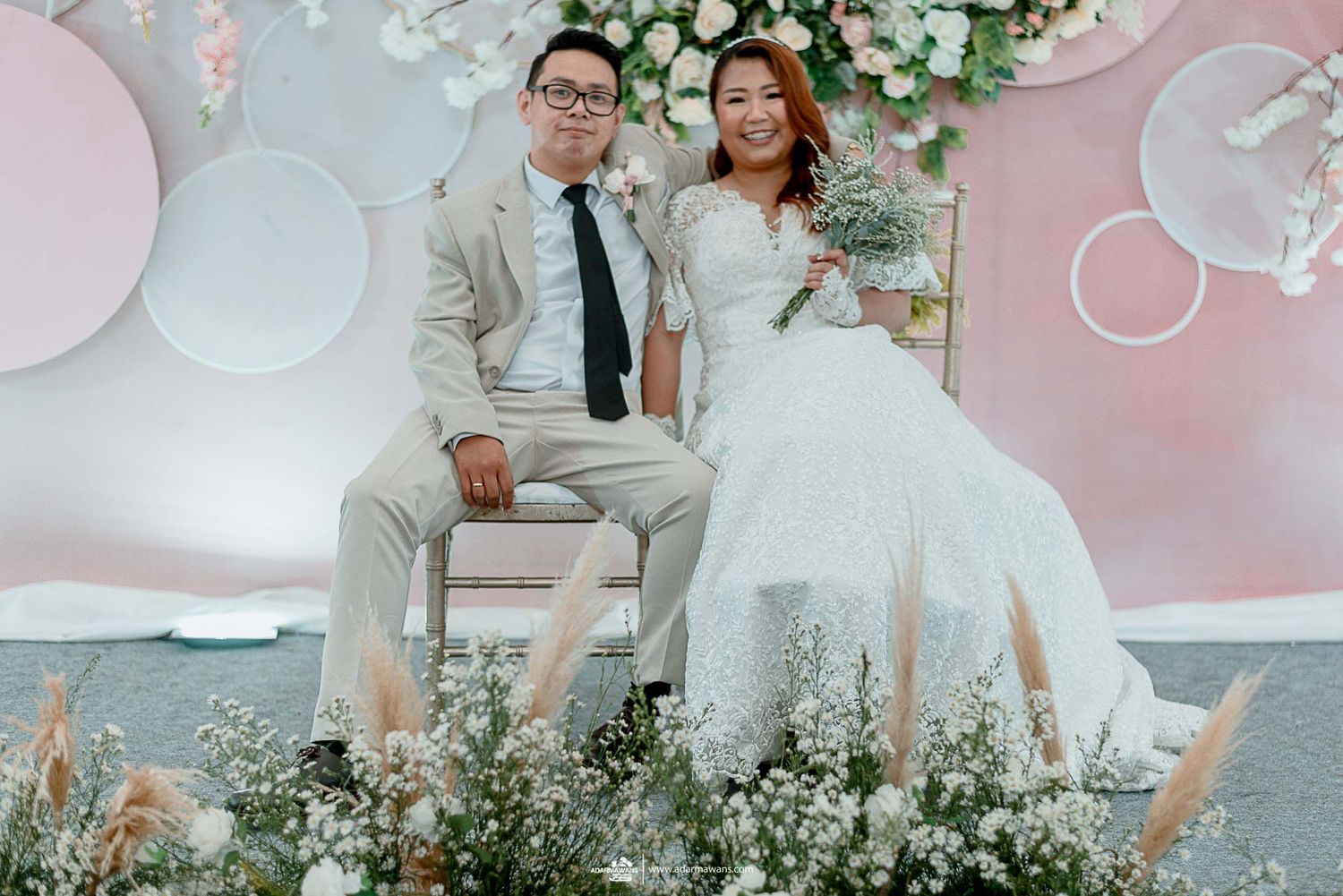 Wedding couple poses on white bench against pink floral backdrop with elegant decorations and flowers.