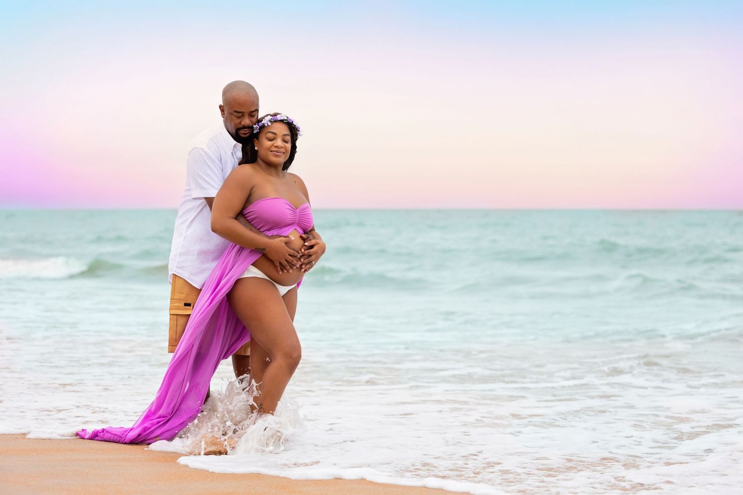 Couple shares intimate moment on beach with flowing pink fabric during sunset photoshoot.