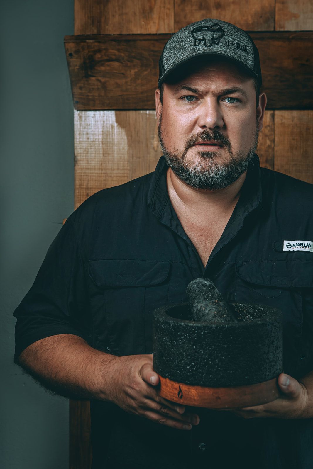 Portrait of a bearded man in a cap holding a black cooking pot.
