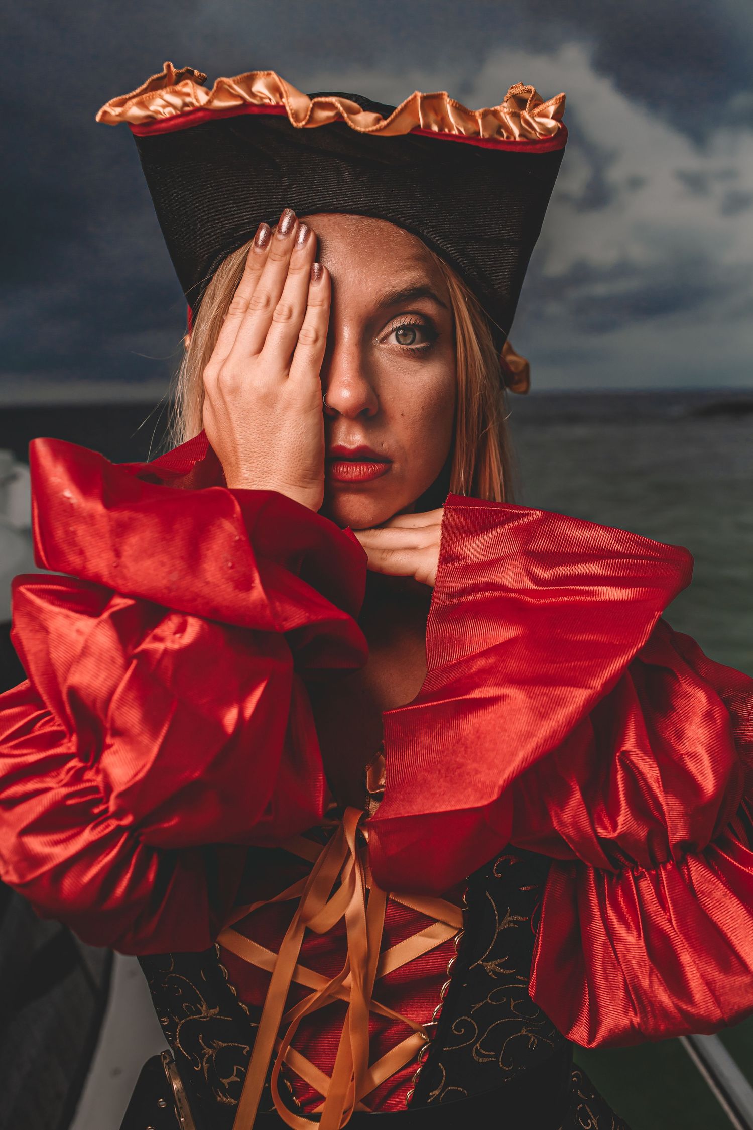 Dramatic portrait in a red ruffled dress and tricorn hat against a moody sky background.