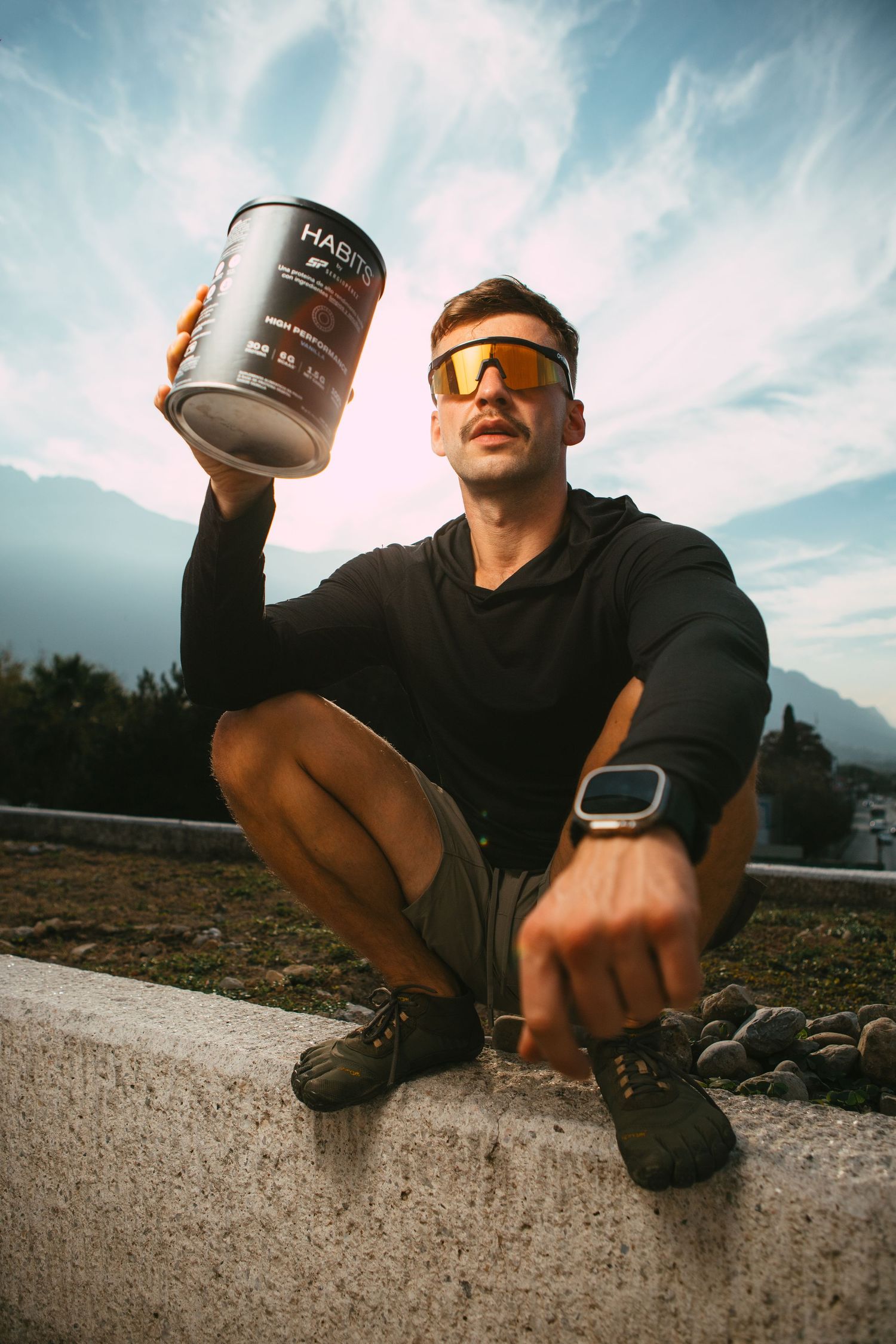 Athletic person in black crouches on concrete holding a large drink container against a mountain backdrop.
